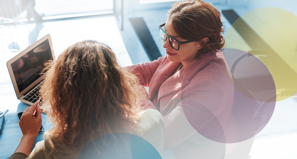 two women converse while sitting in front of an open laptop computer.